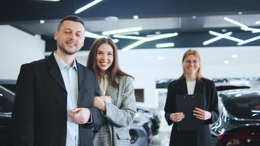 Portrait of happy couple with a saleswoman at an automobile dealership. A manager observes, ready to assist with the deal and paperwork.