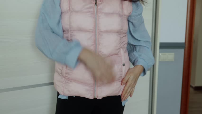 Woman in pink quilted vest and blue shirt smoothing and adjusting vest fabric with both hands in domestic interior.