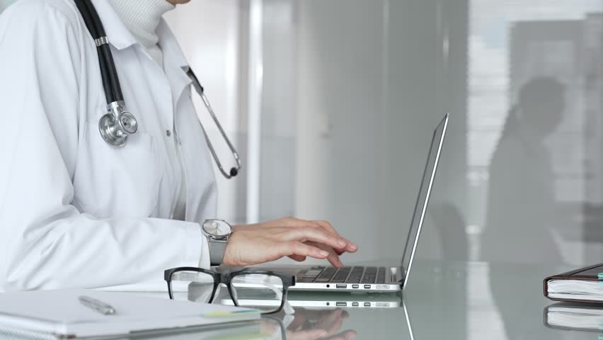 Woman doctor wearing white coat and stethoscope, typing on laptop in clinic office, using modern medical technology. Medicine, healthcare and science concept