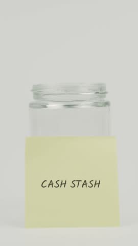 Cash Stash text or word on a yellow sticky note attached to a glass jar with a hand inserting a coin, symbolizing hidden savings, budgeting, and personal money security