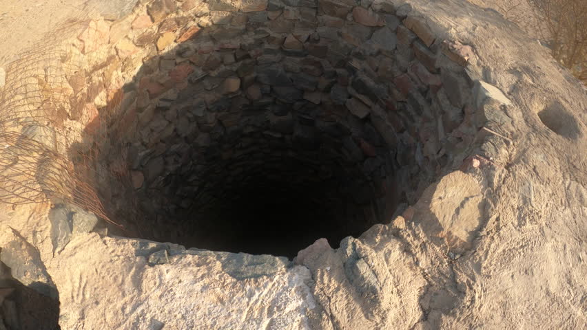 Close-up of an ancient dry-stone well structure with rusted mesh, set against a vast rocky desert and distant mountains under a bright sky — atmospheric B-roll for documentaries, archaeology projects.
