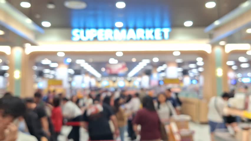 blurry image of a busy interior of a supermarket, the ambiance showing the self-service model in action with numerous people shopping in the aisles.