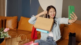 A cheerful young asian woman sits on her comfortable sofa, proudly displays her shopping bags, and happily snaps photos of her colorful haul from a recent shopping trip. - Powered by Shutterstock - Get 15% off with code: PIKWIZARD15