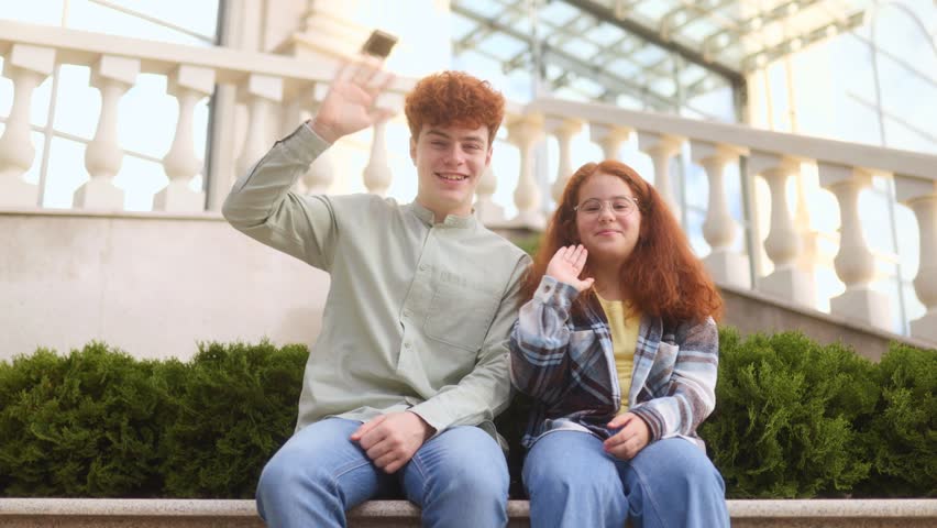 Teenage siblings with red hair wave their hands while sitting outdoors. Casual clothes, natural setting, and simple greeting gesture highlight a lifestyle and youth portrait.