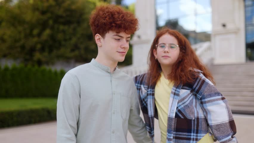Two stylish redhead teenagers, a boy in a shirt and a girl with glasses in plaid jacket, pose outdoors looking directly at the camera in a natural lifestyle portrait.