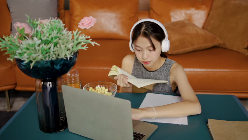 Asian female student enjoys listening to calming music through headphones while engrossed in reading a textbook and and use a laptop computer to find information in her inviting study environment