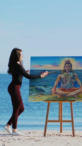 A beautiful woman artist in a tight suit draws a picture. god Shiva is depicted on the picture. summer, near river, on the beach. at sunrise
