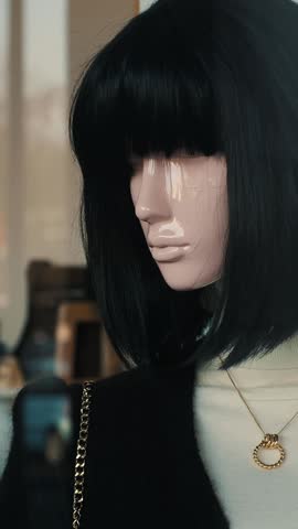 The mannequin is a fair-skinned woman with black hair in a close-up shot. The mannequin is behind glass in a store, wearing a black dress and a necklace with a pendant. The glass reflects the street o