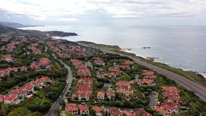 A high aerial pan reveals an affluent coastal community with red roofed houses on green hills, overlooking a highway and the ocean under a cloudy sky.