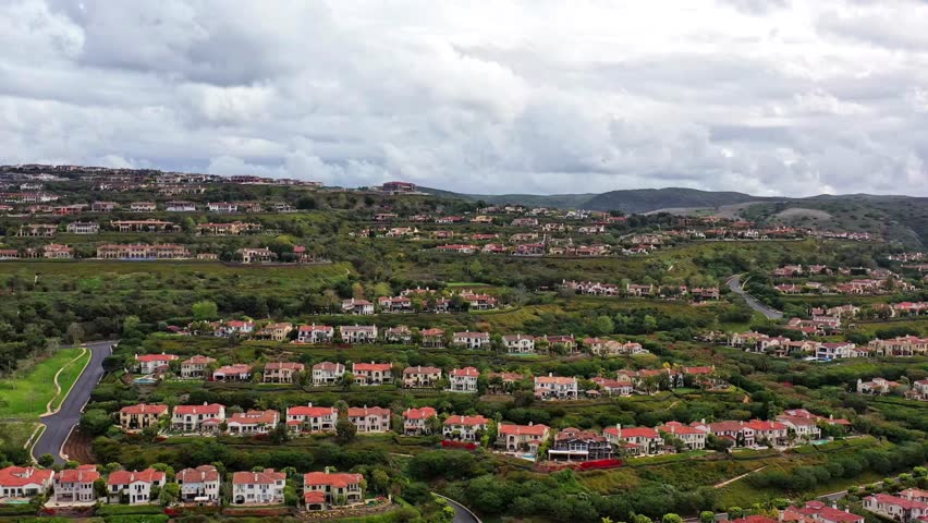 An aerial pan reveals an affluent suburban community with large, red roofed houses nestled in green, rolling hills under a cloudy sky near a highway.