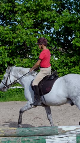 Boy rider, jockey riding on thoroughbred beautiful white stallion, horse, on the training sand field, ground. boy learns to ride a horse in horse riding school. Summer, outdoors