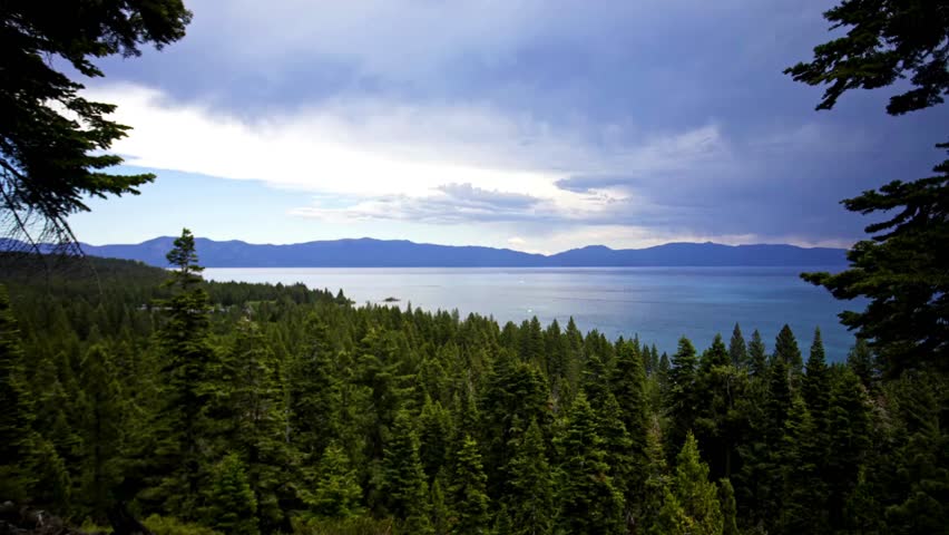 A stunning timelapse captures dramatic clouds and changing light sweeping over a vast pine forest and the beautiful turquoise water of Lake Tahoe until sunset.