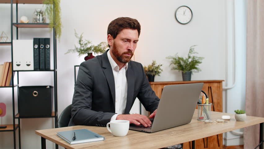 Middle-aged businessman at home office yawns puts head on hands almost asleep then shakes to wake. Freelancer guy at table sips strong coffee straightens and returns to laptop work with fresh energy.