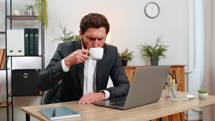 Middle-aged businessman at home office yawns puts head on hands almost asleep then shakes to wake. Freelancer guy at table sips strong coffee straightens and returns to laptop work with fresh energy.