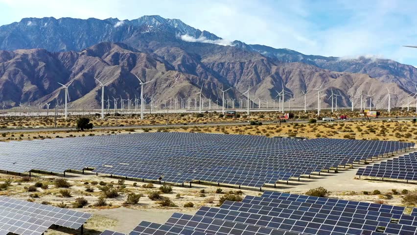 An aerial view shows a large solar farm in the desert foreground, with a busy highway and a vast wind farm set against imposing rocky mountains.