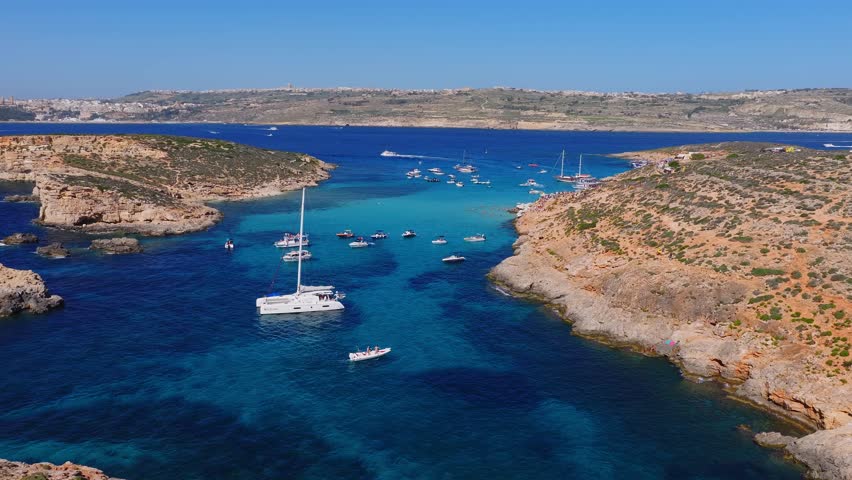 Aerial panorama shows Blue Lagoon on Comino, Malta, with limestone coves, a narrow channel, anchored yachts, fast boats, swimmers, and bright midday light.