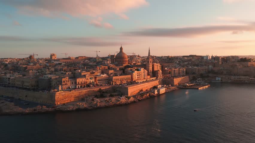 Aerial view of Valletta old town and Grand Harbour at sunset, with basilica dome, cathedral spire, boats and cars in warm light, wide framing and soft backlight.
