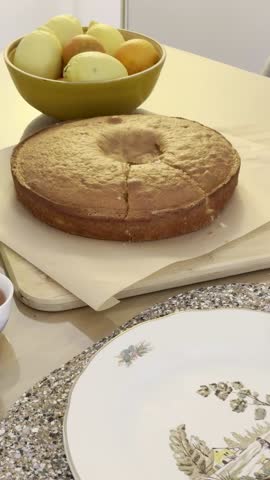 Wide horizontal shot of a hand serving a cake slice onto an ornate plate, emphasizing the modern table setting and tea service elements.