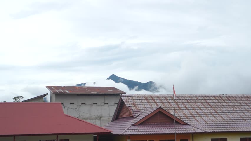 Soft morning fog drifting over green hills with red-roofed houses in the foreground.