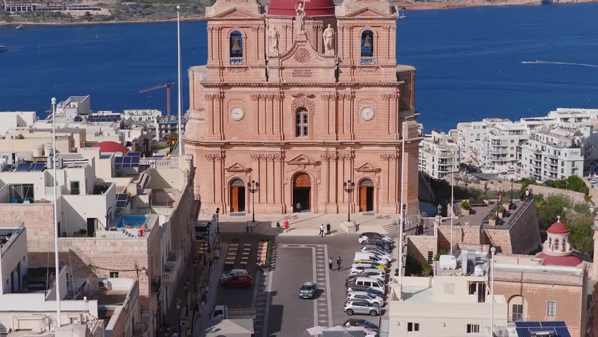 Aerial shot of Mellieha Parish Church, Malta, with red dome and twin towers, plaza activity, boats offshore, Comino and Gozo visible, bright daylight, vivid sea and stone contrast.