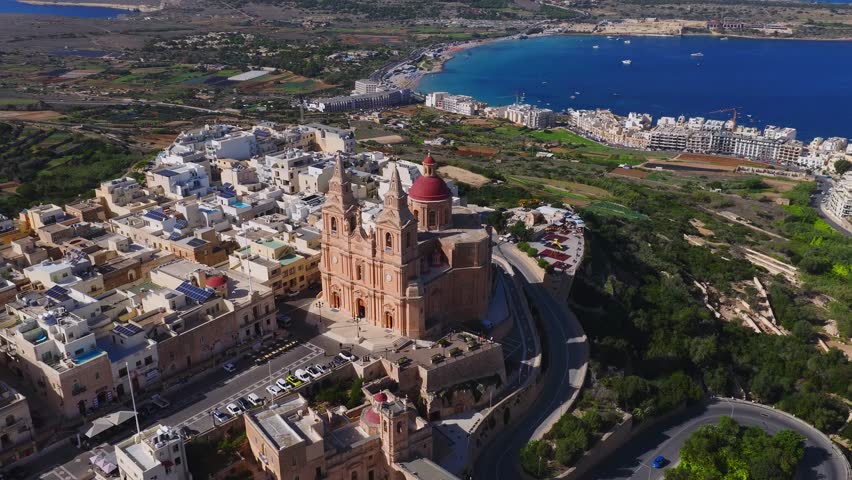 Aerial shot shows Mellieha parish basilica, winding roads, limestone houses, and Ghadira Bay with boats. Panning view includes Comino and Gozo on the horizon.