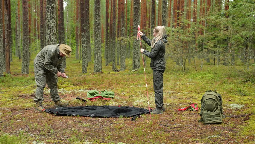 A man and a woman set up a tent in a forest clearing, working close together on stakes and poles. The woman assembles a tent pole while the man prepares ground stakes. Hands and gear are shown in