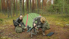 A man and a woman are camping together in a forest with a dog. The travelers are sitting on chairs near a tent. The man is assembling a portable camping table. - Powered by Shutterstock - Get 15% off with code: PIKWIZARD15