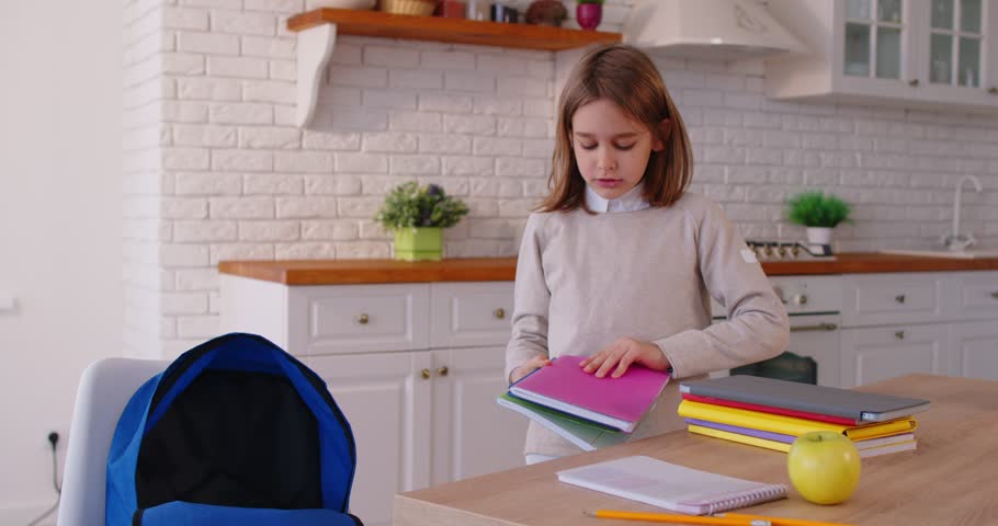 Cheerful school child packing colorful notebooks into backpack on kitchen table, preparing for school day. Focused young pupil organizing study materials and supplies before going to school.