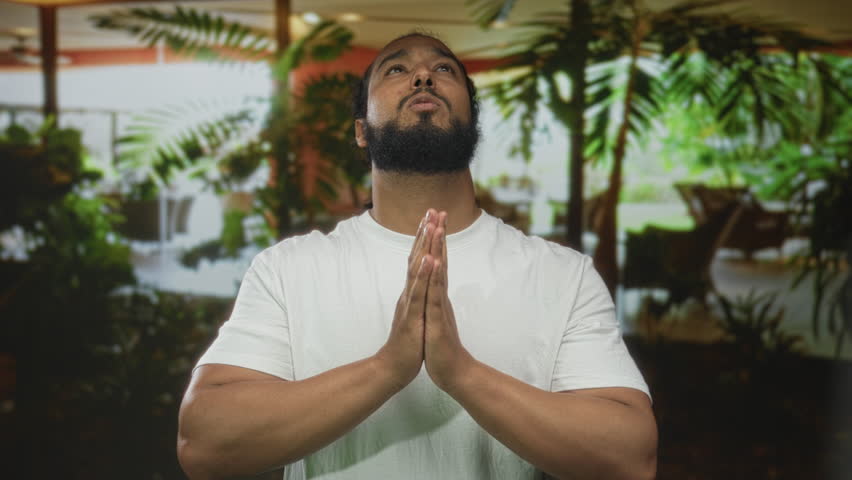 Man praying with hands pressed together in hotel building lobby amid indoor plants and seating; serenity gratitude reflection.