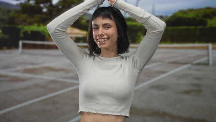 Hispanic woman raises arms overhead on outdoor tennis court in sportswear with net and greenery behind; joy.