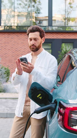 Handsome Caucasian man connecting a car charger to his electric vehicle while using a mobile app on his smartphone to start the charging process