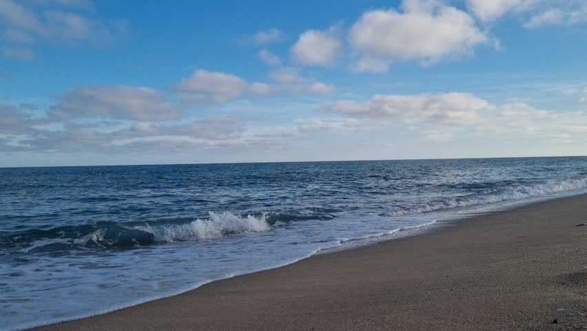 Waves gently wash onto a dark sandy beach under a sky filled with fluffy clouds. Ideal for backgrounds, nature scenes, travel themes, or weather footage.