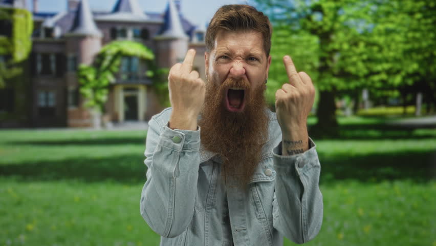 Man with full red beard showing middle fingers while shouting with open mouth in a sunlit green park; anger.