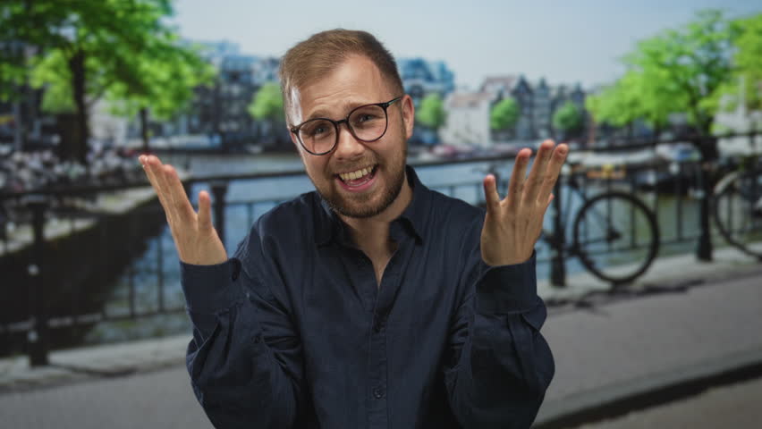 Man wearing glasses and navy shirt clenches fists and smiles on street by amsterdam canal near bicycle railing; victory joy.
