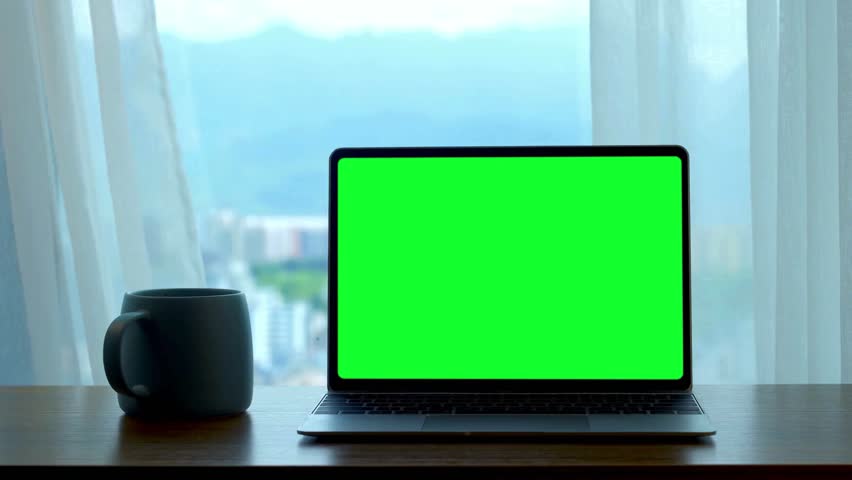 A laptop with a green screen sits on a wooden table next to a mug, with a window and white curtains in the background. Perfect for virtual backgrounds and mockups.