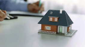 A miniature house sits on a white table while a person writes in the background. This image represents real estate, property investment, or home ownership planning. - Powered by Shutterstock - Get 15% off with code: PIKWIZARD15