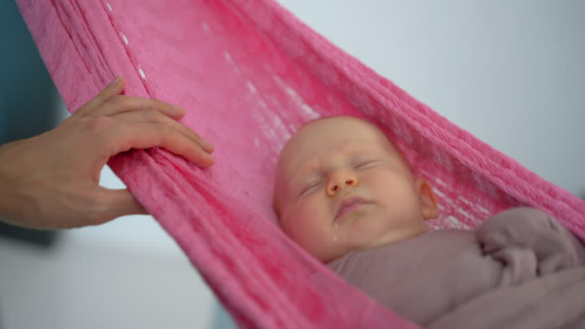 Loving father gently swings his swaddled two-month-old baby in a hammock to help him fall asleep, creating a peaceful and nurturing family bedtime moment.
