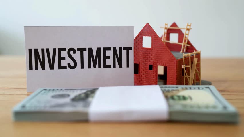 A close-up shows an investment sign, a house model, and a stack of money on a wooden surface, set indoors in a bright setting, symbolizing financial growth.