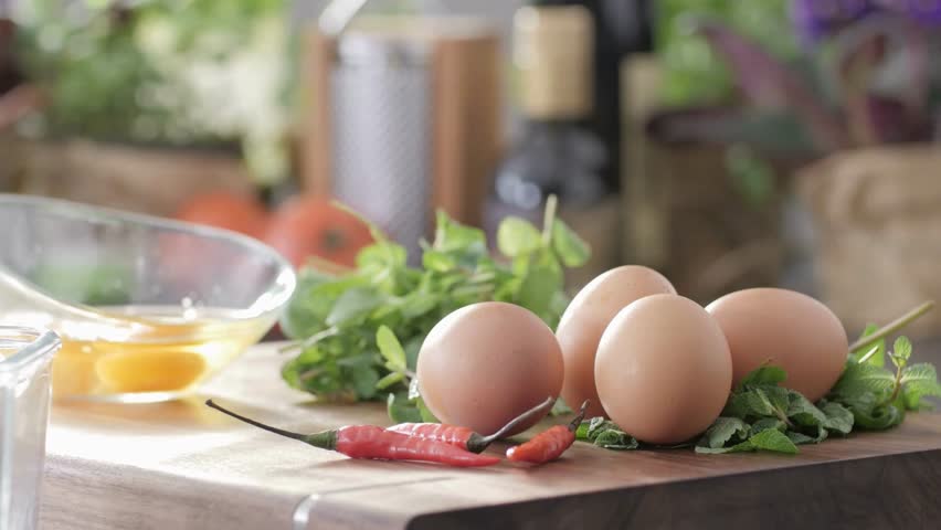 a close up view shows eggs and herbs on a wooden board, with a bowl of yolks in the background. a chili pepper adds a touch of spice to the scene, hinting at a delicious meal.