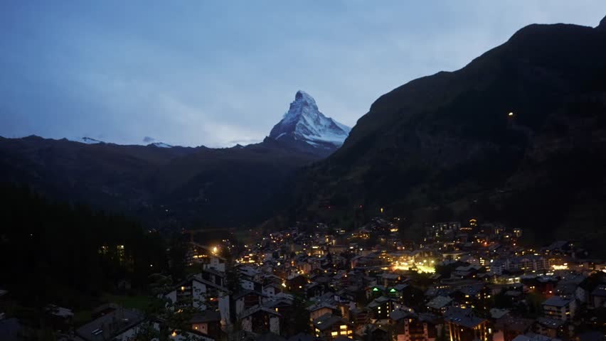 A young man walks across the frame overlooking Zermatt at blue hour, with the Matterhorn mountain in the distance. Scenic travel footage showing the illuminated alpine town and mountain landscape