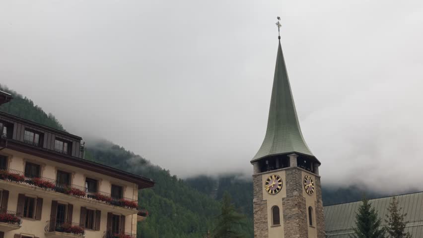Close-up shot of the church tower in Zermatt, Switzerland, on a cloudy day. Architectural detail with mist-covered mountains in the background, capturing alpine atmosphere and culture