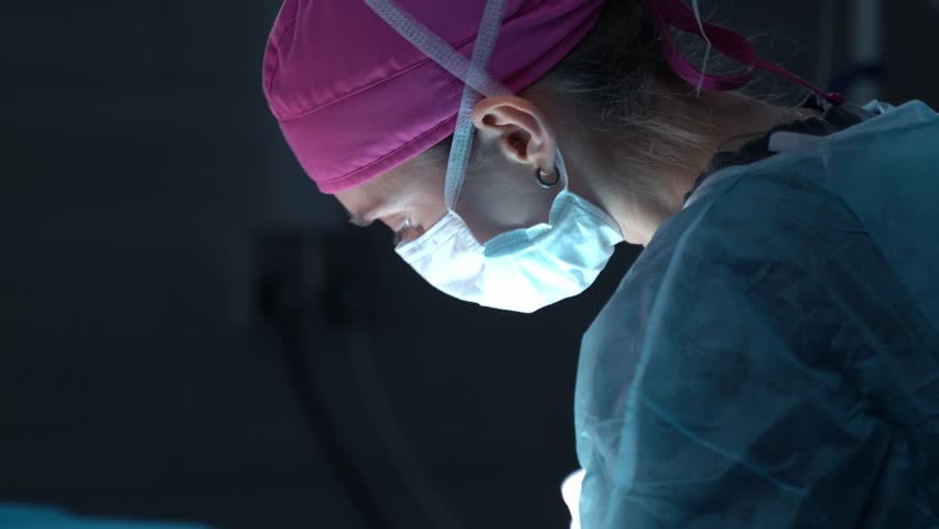 Young female surgeon in a sterile gown and mask concentrating on a patient during a medical procedure under the bright light of a surgical lamp in a dark operating room - Powered by Shutterstock - Get 15% off with code: PIKWIZARD15
