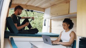 Young diverse couple enjoying van life together in nature. Woman working remotely on a laptop while her partner relaxes using a tablet on the bed - Powered by Shutterstock - Get 15% off with code: PIKWIZARD15
