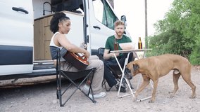 Interracial couple relaxing in nature next to their camper van. Woman playing the guitar and petting their boxer dog while her boyfriend snacks - Powered by Shutterstock - Get 15% off with code: PIKWIZARD15