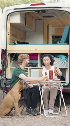 Happy diverse couple with their dog enjoying a meal and beer on a camping trip. Friends celebrating their holiday with a converted van life