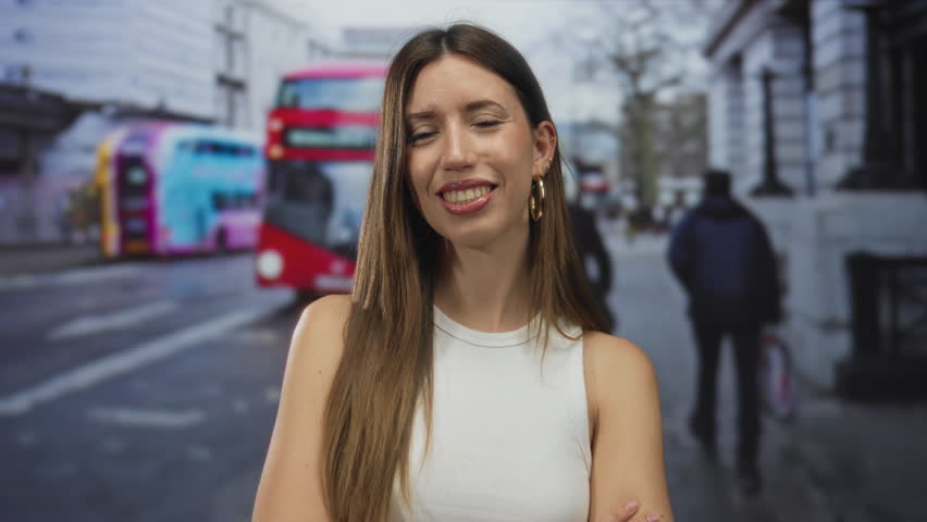 Woman with crossed arms next to red double decker bus on city street, wearing white tank top and hoop earrings; confidence.