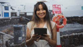 Woman brunette holding tablet with both hands smiling on street by seaside pier and lifebuoy, young latina in sleeveless top; serenity connection. - Powered by Shutterstock - Get 15% off with code: PIKWIZARD15