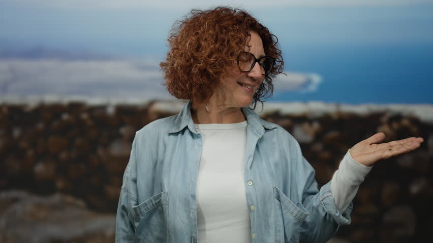 Woman extends hand by seaside promenade with relaxed expression, wearing casual attire and smiling, against a backdrop of sea and horizon, showcasing a serene outdoor moment.