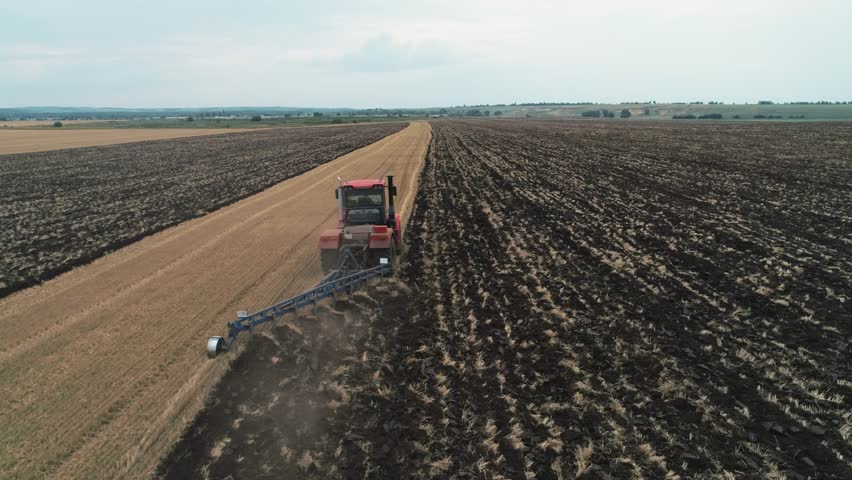 Tractor plowing field across farmland turning soil into dark furrow under cloudy sky with tractor towing plow while harvest stubble and crop residue line tractor path in expansive rural land