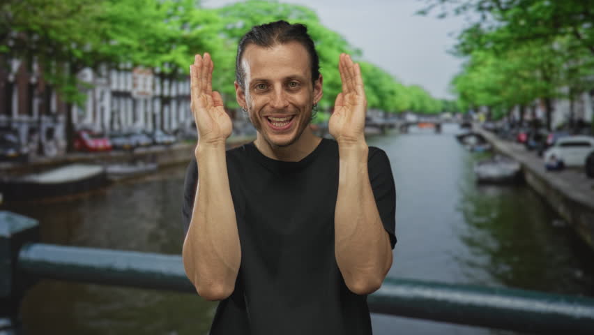 Young hispanic man with long hair covering his face with hands on an amsterdam canal street wearing black t shirt and smiling; joy travel memory.